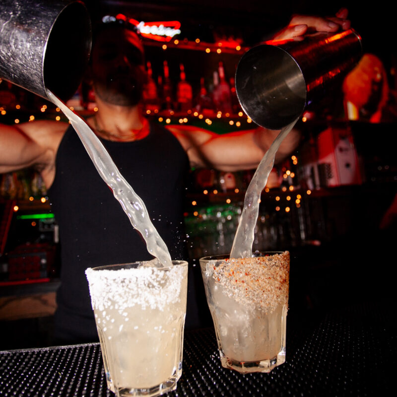 A bartender at Warehouse pours two cocktails simultaneously into salt-rimmed glasses, capturing the vibrant energy of the bar.
