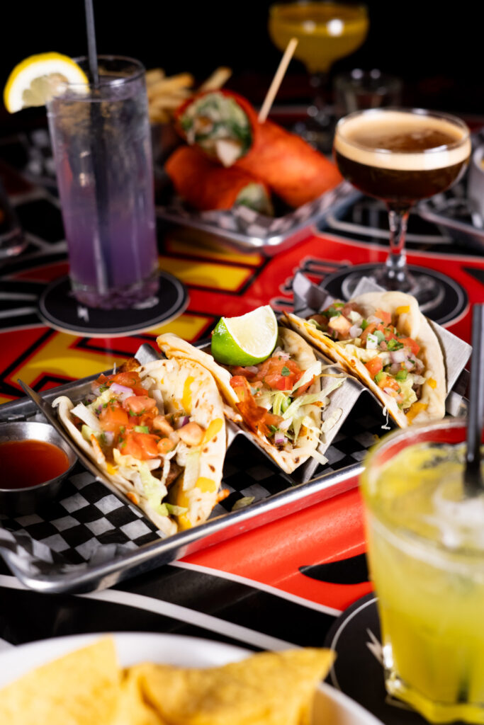 Three street tacos in a plate, surrounded by colorful cocktails and other dishes.