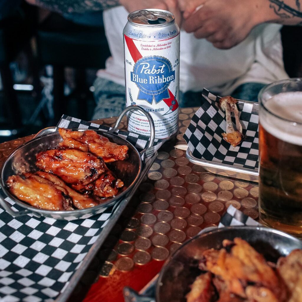Glazed chicken wings served with Pabst Blue Ribbon beer at El Furniture Warehouse on a penny-covered tabletop.