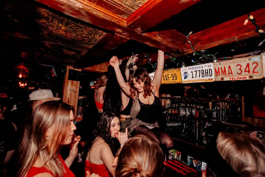 Group of friends dancing on the bar at Warehouse with arms raised while bottles line the back counter.