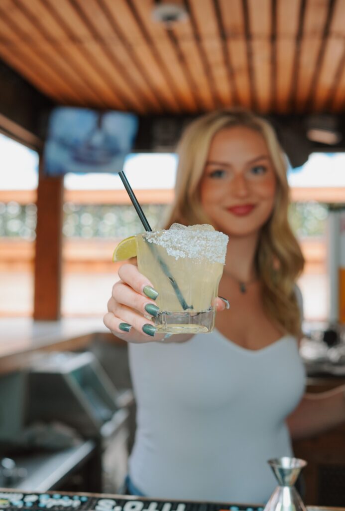 A woman with a welcoming smile holding out a salt-rimmed margarita.