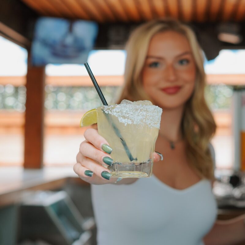 A woman with a welcoming smile holding out a salt-rimmed margarita.