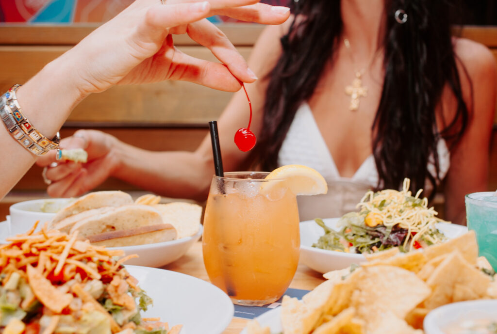 Close-up of a hand holding a maraschino cherry above an orange cocktail glass at Warehouse, ready to garnish the drink.