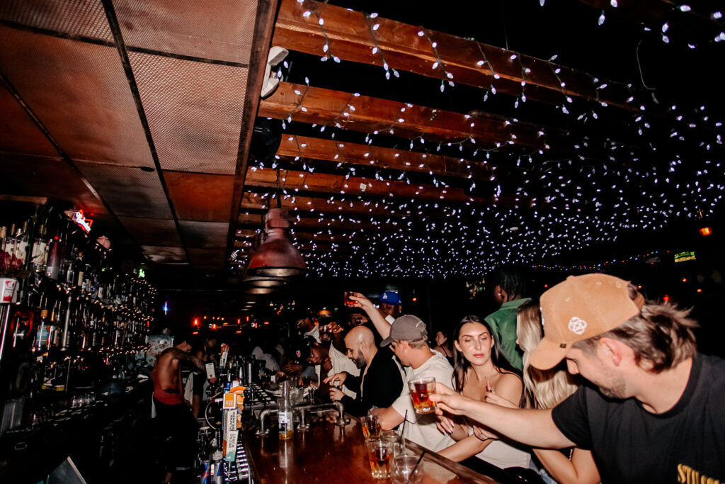 Night scene at Warehouse with guests standing and drinking around a wooden bar counter.