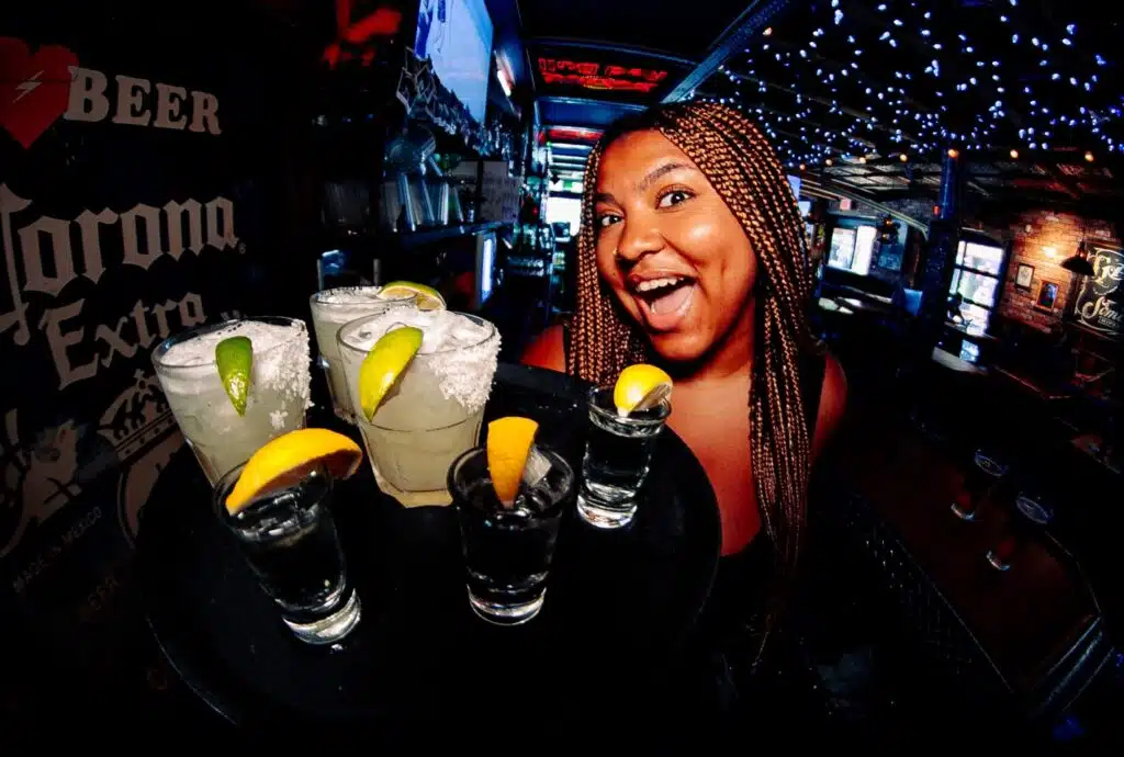 Smiling woman in a busy bar holding a tray of colorful shot glasses garnished with citrus.