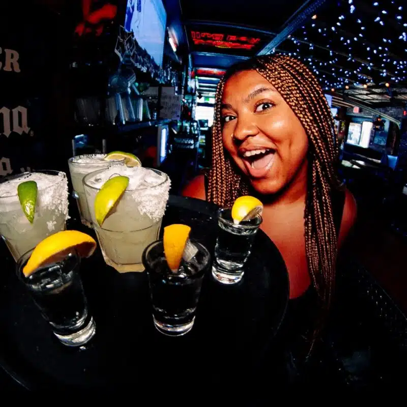 Smiling woman in a busy bar holding a tray of colorful shot glasses garnished with citrus.