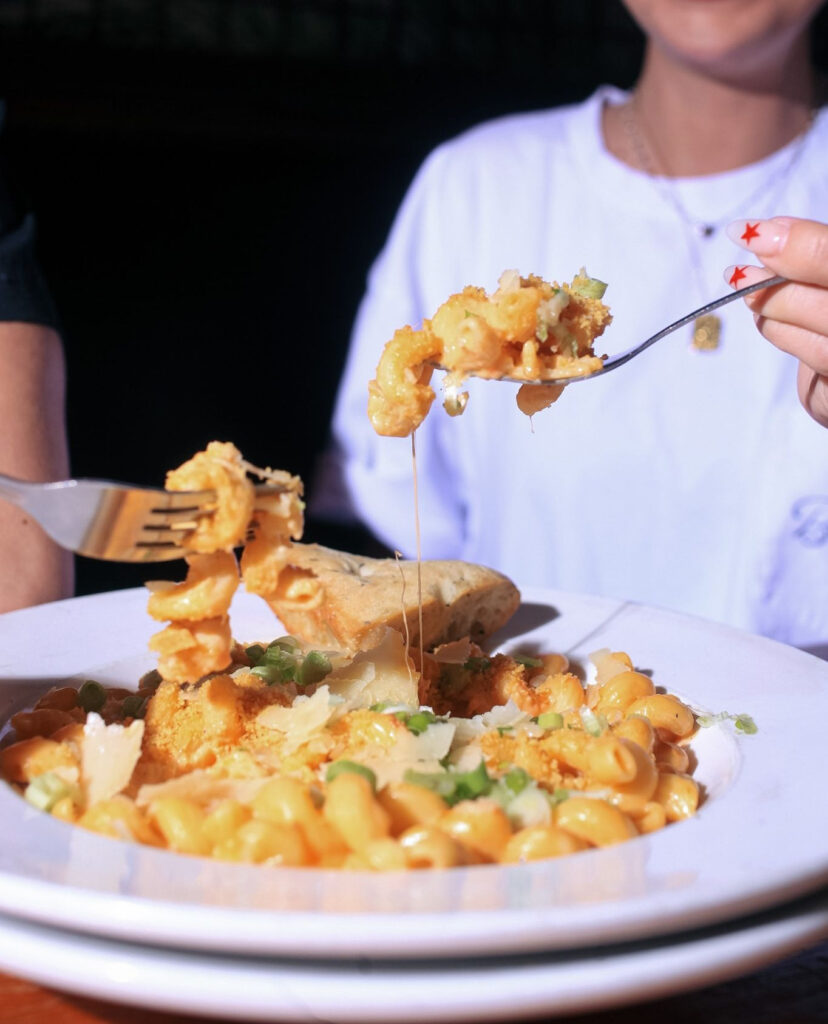 Close-up of Warehouse’s mac and cheese being shared, featuring gooey cheese, green onions, and garlic bread.