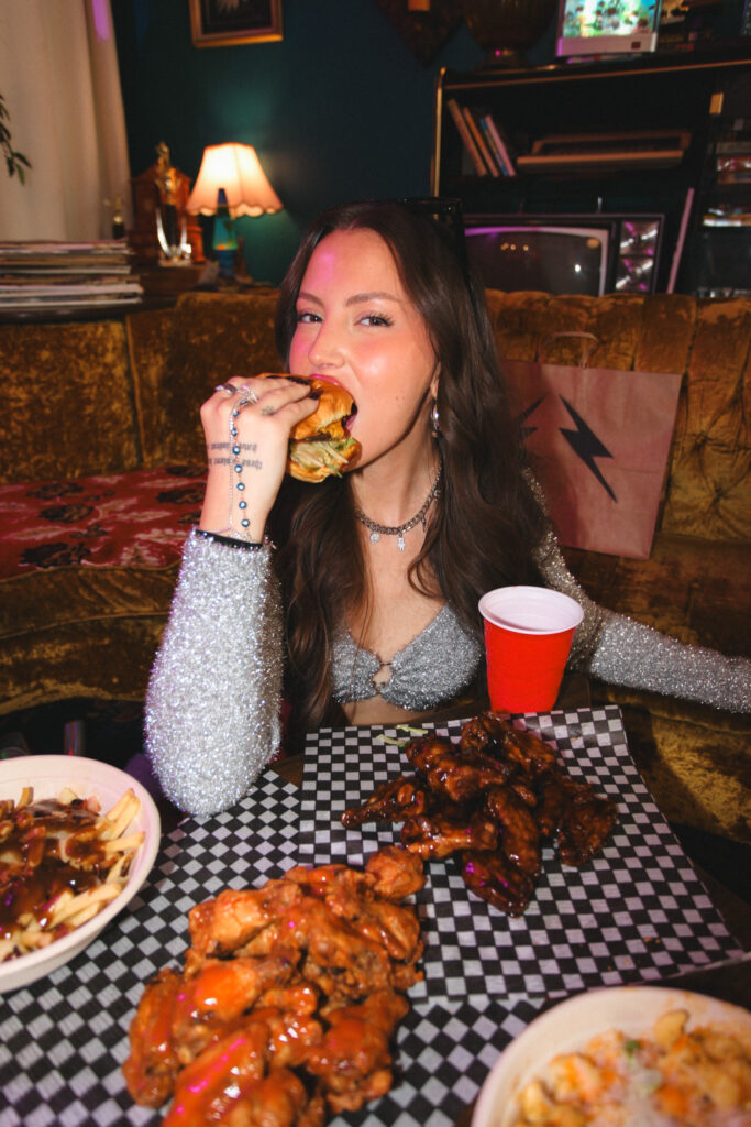 A young woman taking a big bite of a burger while sitting at a table covered with plates of chicken wings and fries.