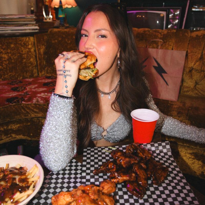 A young woman taking a big bite of a burger while sitting at a table covered with plates of chicken wings and fries.