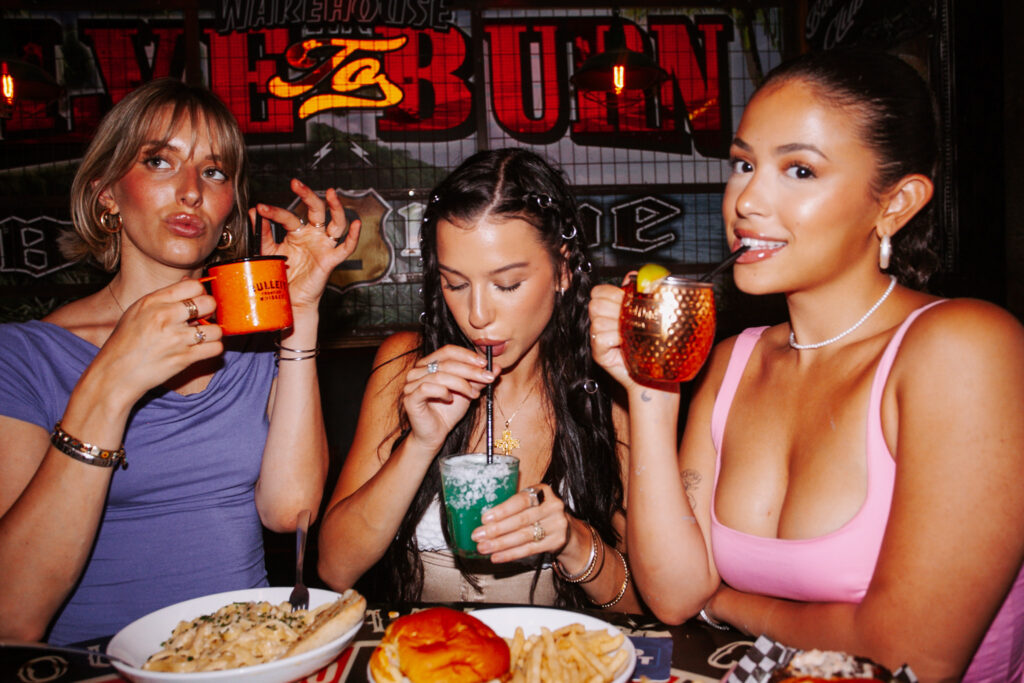 Three women smiling and posing around a table at Warehouse filled with comfort food and drinks, including a Moscow Mule.