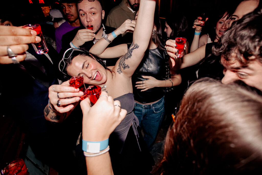 A group of young adults laughing and raising red shot glasses for a toast in a crowded, dimly lit bar.