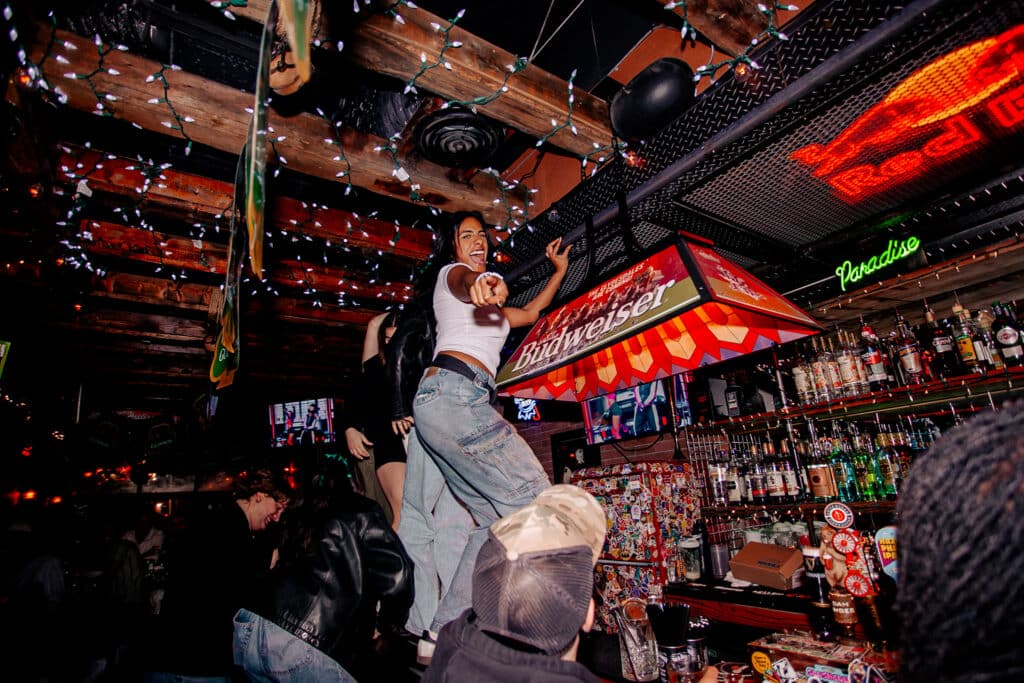 A woman in jeans and a white top cheers while standing on a table inside Warehouse, decorated with string lights.