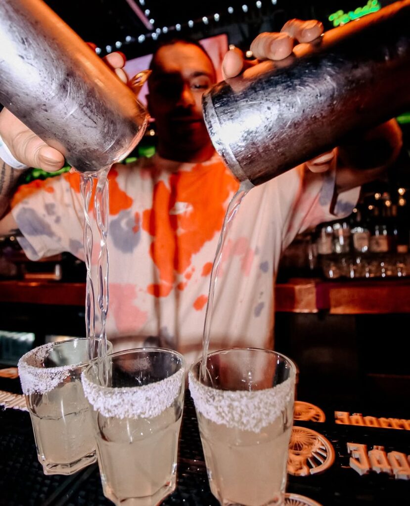 A bartender at Warehouse pours cocktails from metal shakers into salt-rimmed glasses behind the bar.