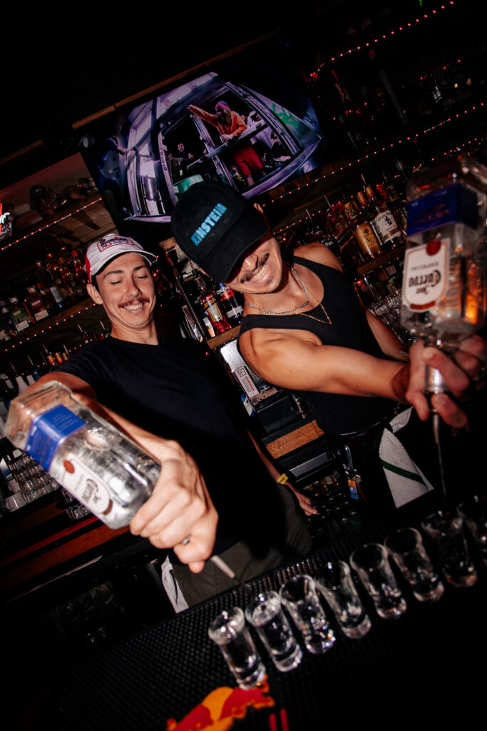 Bartenders at Warehouse pouring vodka into shot glasses behind a lively bar during a World Cup watch party.