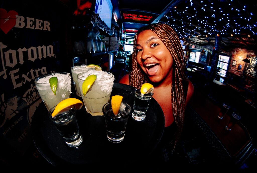Smiling woman holding a tray of drink shots with citrus, in a bar.