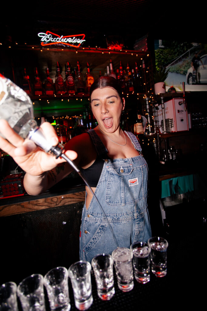 Warehouse bartender pouring and serving vodka shots during a busy late-night gathering.