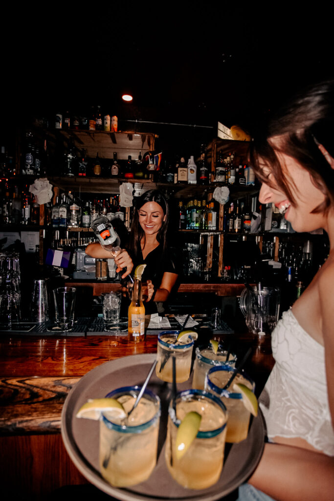 Two Warehouse team members laughing while serving drinks and interacting with customers in a welcoming restaurant atmosphere.